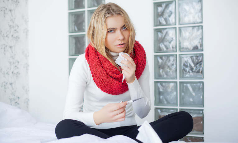 mladá žena se šálou kolem krku sedí na posteli a v rukou drží teploměr a ubrousek