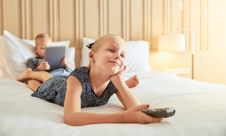 holčička leží na břiše na manželské posteli a v ruce drží dálkové ovládání k televizi
