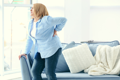Žena trpí bolestí spodní části zad a opírá se proto o opěradlo gauče při vstávání